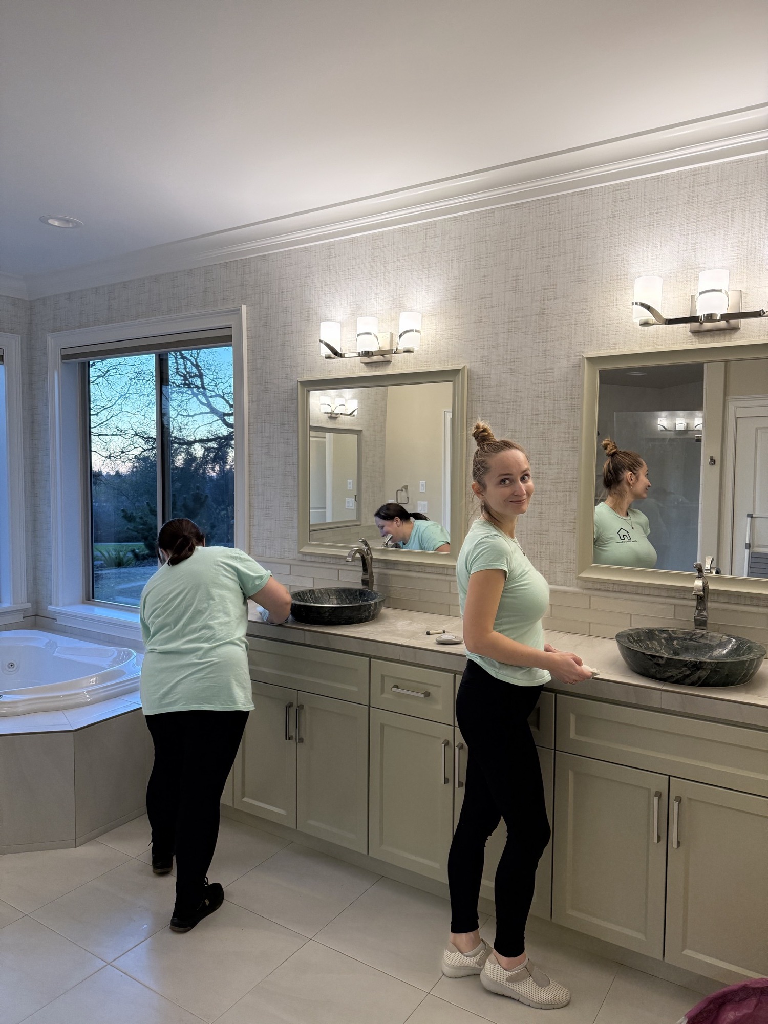 Two cleaners cleaning a primary bathroom vanity, one visible in the mirror